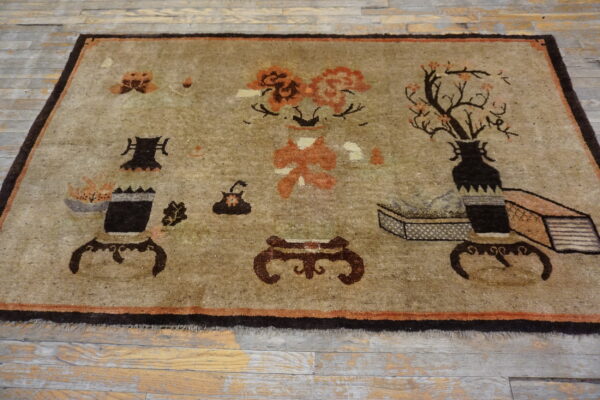 Muted tan rug with dark brown borders and stylized antique object patterns resting on worn wood flooring.