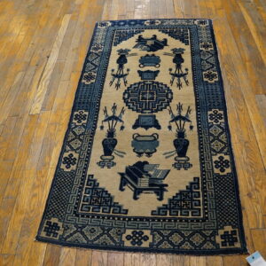 Low-pile tan rug with dark blue traditional patterns, including vases and geometric shapes, on a worn wood floor.