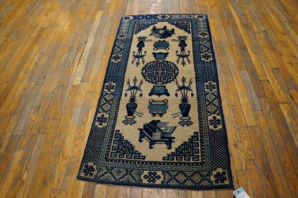 Low-pile tan rug with dark blue traditional patterns, including vases and geometric shapes, on a worn wood floor.