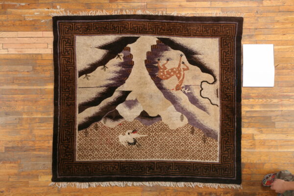 Pictorial rug featuring deer, birds, and mountains surrounded by a dark brown geometric border on wood.