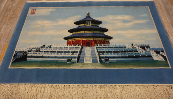 Blue pictorial rug depicting a tiered temple structure on a platform with cream fringe on wood floor.