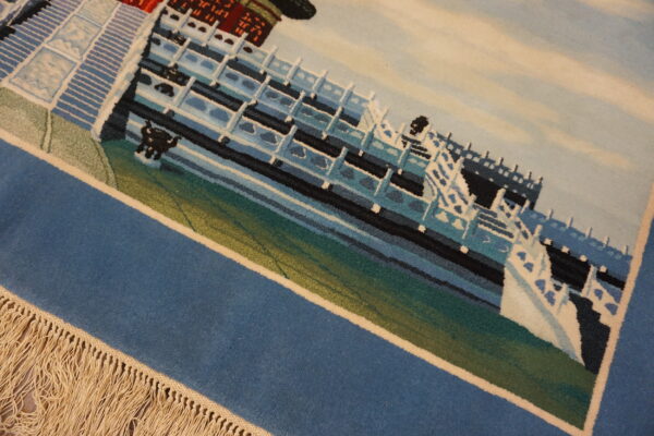 Close-up of a low-pile rug with a detailed scenic pattern in blue, white, teal, and green, bordered by fringe.