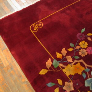 A plush maroon rug with a multicolored floral pattern and gold border sits on light hardwood flooring.