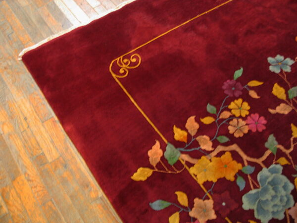 A plush maroon rug with a multicolored floral pattern and gold border sits on light hardwood flooring.