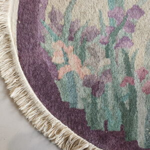 Round high-pile rug detail shows purple, green, and pink flowers, cream fringe, and light tiled flooring.