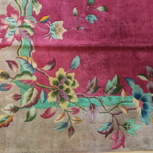 Detailed view of a deep pink rug with a colorful, elaborate floral border on a textured tan background.