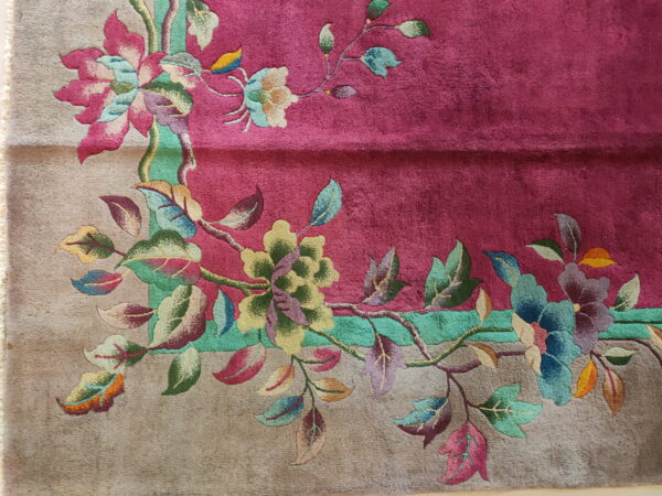 Detailed view of a deep pink rug with a colorful, elaborate floral border on a textured tan background.