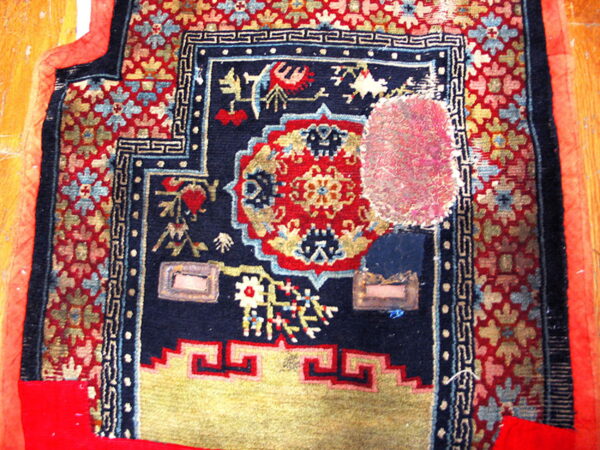 Low-pile rug fragment in navy, red, and gold, showing complex patterns, wear, and a bright pink patch.