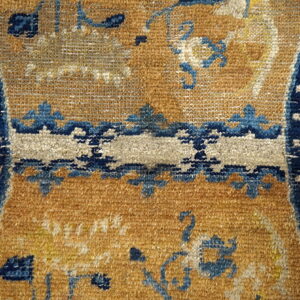Textured rug detail with a golden-brown field, indigo borders, and stylized blue and cream motifs.