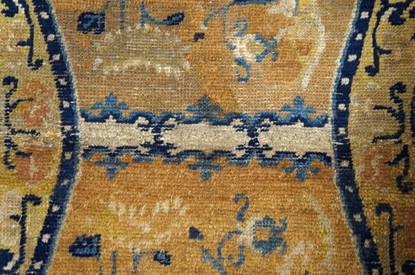 Textured rug detail with a golden-brown field, indigo borders, and stylized blue and cream motifs.