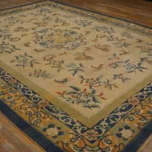 A beige floral rug featuring blue, gold, and green borders is displayed on rustic wooden floorboards.
