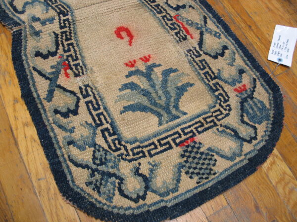 Tan low-pile rug with navy blue fretwork borders and central blue and red floral motifs on wood flooring.