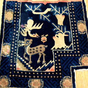 Dark blue rug with tan figures of deer and animals, detailed borders, resting on light wood floors.