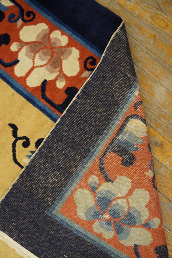 Close-up of a yellow and orange-red floral rug, folded back to show the navy woven backing on wooden floorboards.