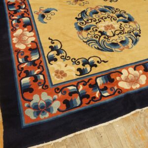 Gold and navy rug with orange floral border resting on distressed yellowish wood plank flooring.