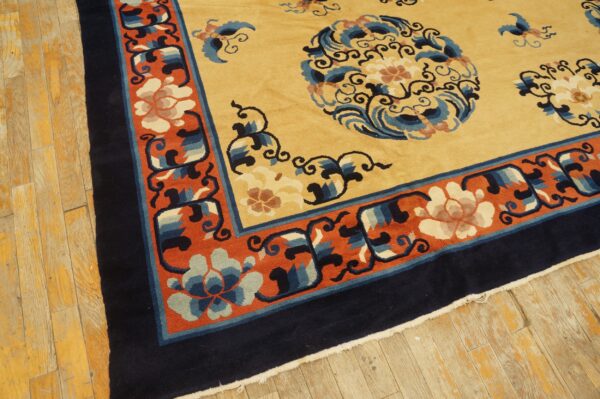 Gold and navy rug with orange floral border resting on distressed yellowish wood plank flooring.