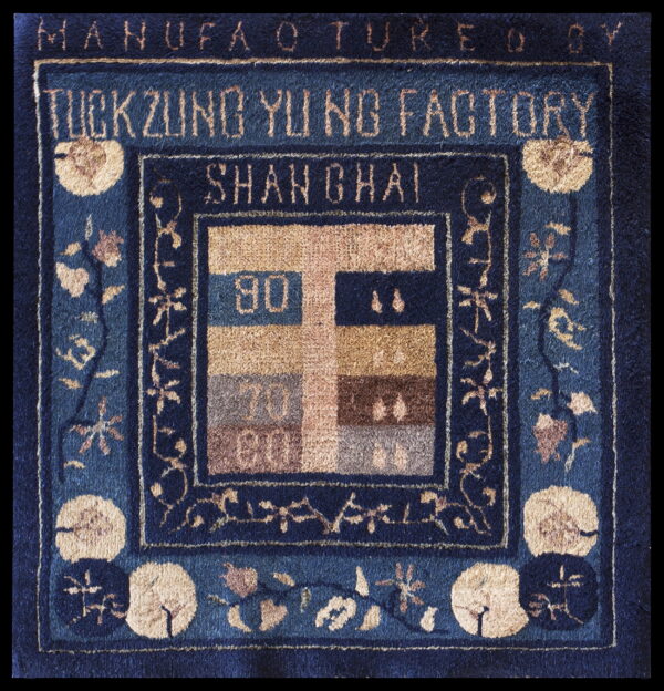 Close-up of a blue rug with tan floral motifs framing factory text and a numbered color block design.
