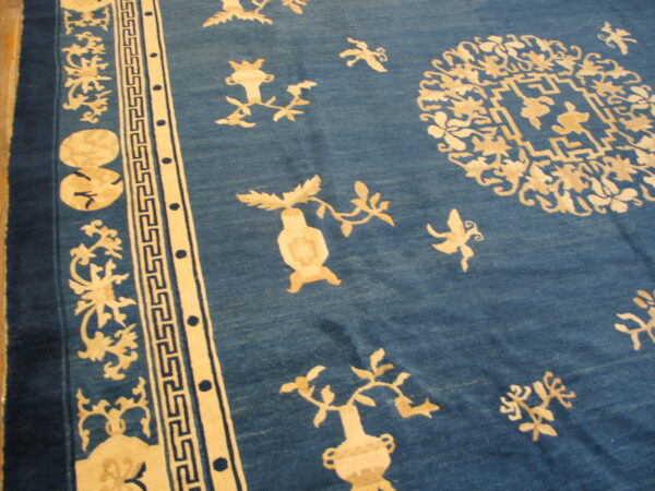 Blue rug featuring scattered light tan floral and vase motifs bordered by a geometric fret pattern.