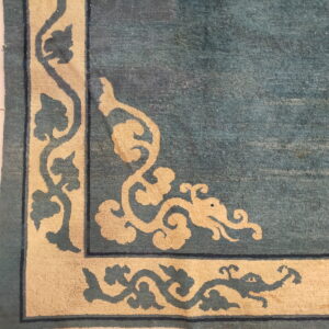 Corner detail of a blue-green rug featuring a thick cream scrolling border, resting on white square tiled flooring.