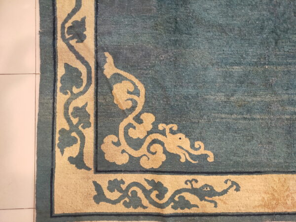 Corner detail of a blue-green rug featuring a thick cream scrolling border, resting on white square tiled flooring.