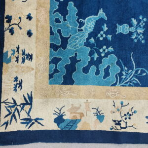 Corner of a blue and cream rug featuring stylized birds and bamboo, resting on gray concrete.