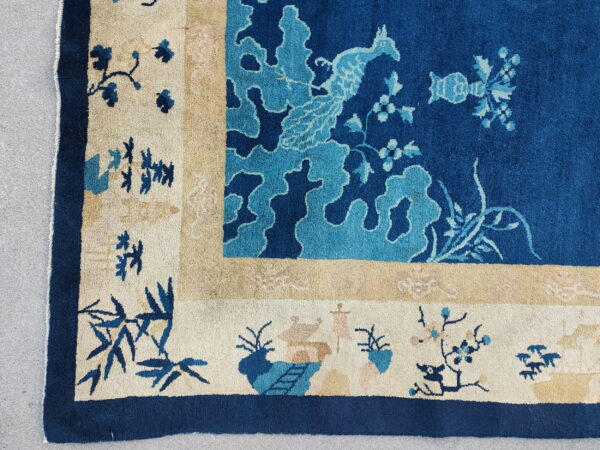 Corner of a blue and cream rug featuring stylized birds and bamboo, resting on gray concrete.
