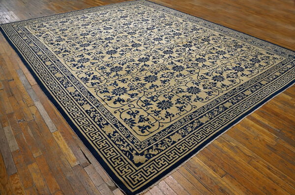 Tan rug with dense blue floral patterns and broad geometric fretwork borders on wood planks.