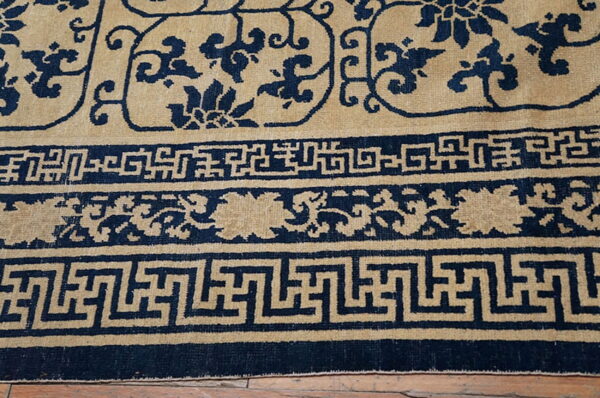 Tan and navy low pile rug featuring a scrolling floral field and detailed geometric border patterns.