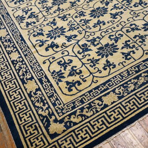 A tan and navy rug with floral motifs and geometric borders rests on rustic hardwood floors.
