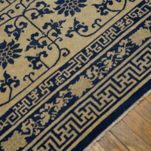 Low-pile rug in tan and navy blue displaying floral patterns and a complex geometric border on wood flooring.