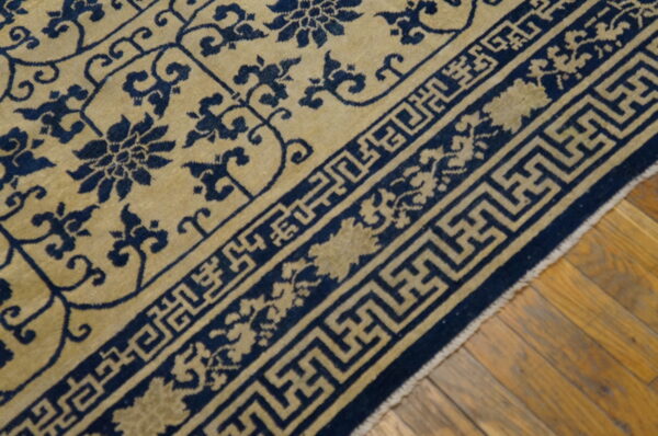 Low-pile rug in tan and navy blue displaying floral patterns and a complex geometric border on wood flooring.