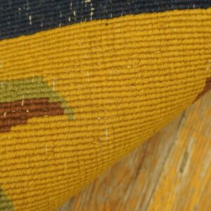 Detail shot of a flatweave rug curled on a distressed yellow floor, featuring navy, mustard, and earthy tones.