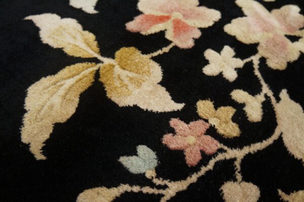Close-up detail of a dense black rug featuring pink, gold, and cream floral and leaf shapes in cut pile.