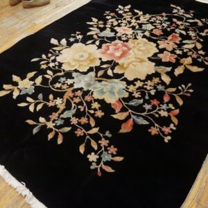 Black high-pile rug featuring large floral motifs in cream, pink, and blue on worn wooden floorboards.