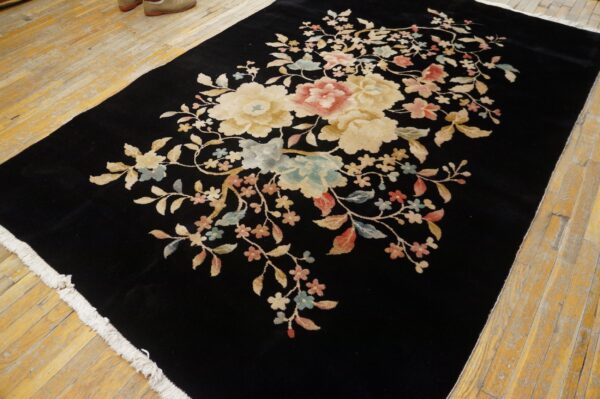 Black high-pile rug featuring large floral motifs in cream, pink, and blue on worn wooden floorboards.