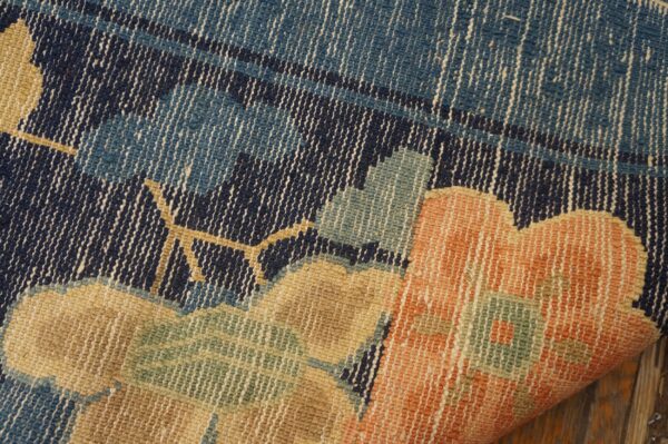 Woven rug close-up showing orange, tan, blue, and green floral patterns on navy, curling over wood planks.