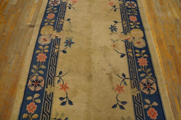Tan runner rug featuring wide blue borders with floral and geometric designs rests on distressed wood flooring.