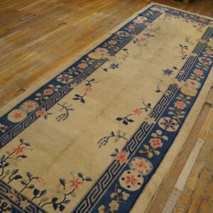 Tan runner rug with a navy floral and key-pattern border rests on worn hardwood planks.