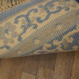 A rolled-up blue rug with a light beige geometric border and fringe lies on brown wood plank flooring.