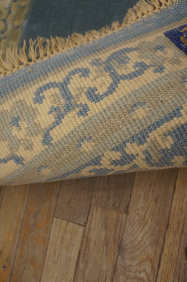 A rolled-up blue rug with a light beige geometric border and fringe lies on brown wood plank flooring.