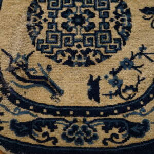 Tan rug with dark blue traditional patterns, including a geometric medallion and stylized figures, sitting on wood flooring.