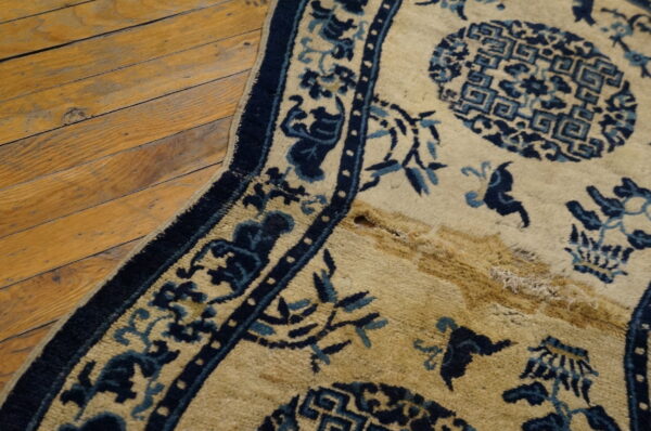 A beige and blue patterned rug with a damaged section exposing shaggy pile lies on a wood floor.