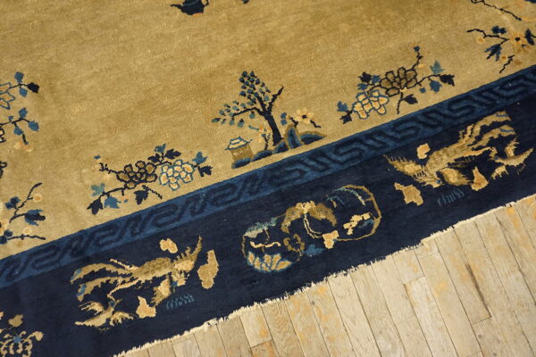Tan rug field and navy border featuring blue and gold floral and figurative patterns resting on wood planks.