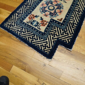 A cream and blue rug with floral patterns rests on a hardwood floor next to a black shoe.