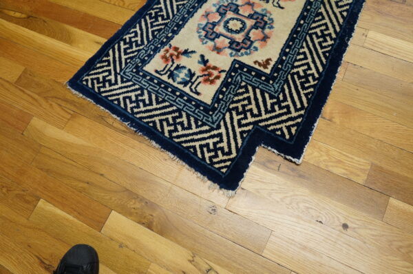A cream and blue rug with floral patterns rests on a hardwood floor next to a black shoe.