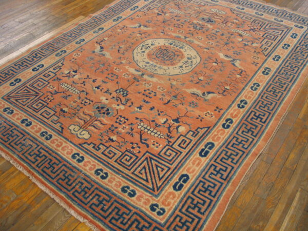 Low-pile rug with salmon field, blue fretwork borders, and floral patterns on wood flooring.