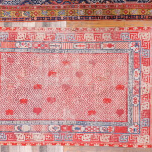 Low-pile red rug featuring a tight cream geometric pattern and blue borders rests on a wooden floor.
