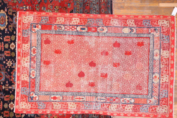 Red and cream rug featuring a dense geometric fretwork pattern and complex blue borders on wood flooring.