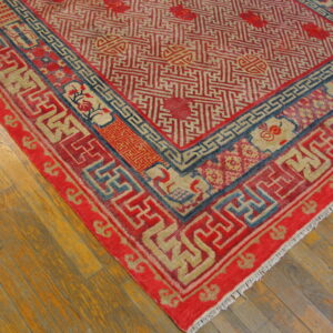 Intricate red and beige fretwork rug with dark blue accents rests on honey-toned wood planks.