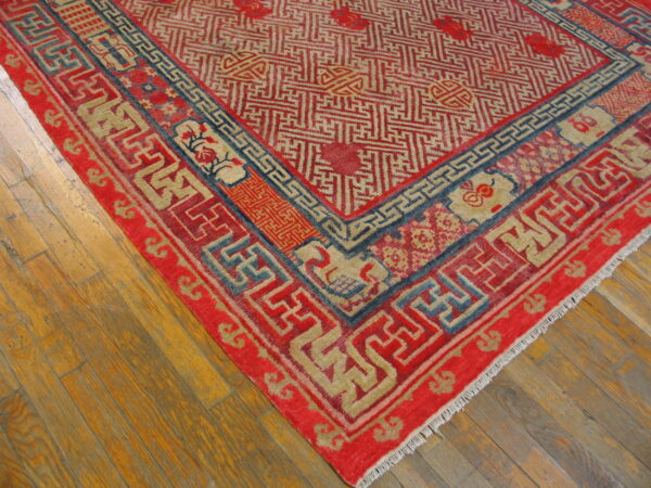 Intricate red and beige fretwork rug with dark blue accents rests on honey-toned wood planks.